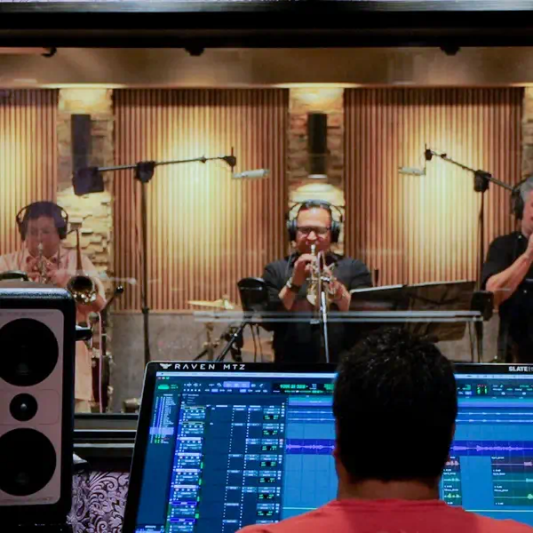 Musicians performing in a recording studio while a technician handles a mixer console outside.